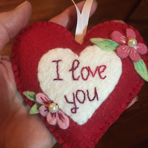 Może przedstawiać: Czerwona filcowa ozdoba w kształcie serca z białym środkiem. Słowa "I love you" są wyhaftowane na czerwono. R&oacute;żowe filcowe kwiaty z perłowymi akcentami i zielonymi liśćmi zdobią serce. Dołączona jest biała wstążka.