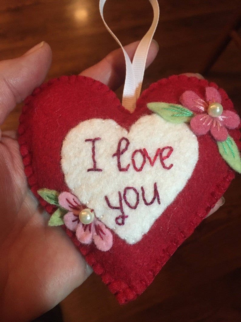 Może przedstawiać: Czerwona filcowa ozdoba w kształcie serca z białym środkiem. Słowa "I love you" są wyhaftowane na czerwono. R&oacute;żowe filcowe kwiaty z perłowymi akcentami i zielonymi liśćmi zdobią serce. Dołączona jest biała wstążka.