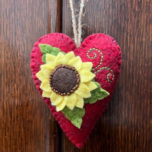 May include: A heart-shaped, red felt ornament with a sunflower applique. The sunflower has yellow petals and a brown center. Green felt leaves and embroidered green vines accent the design. A twine loop is attached for hanging.