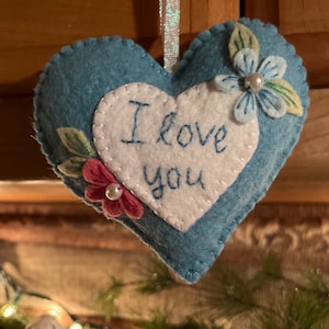 Może przedstawiać: Niebieska filcowa ozdoba w kształcie serca z białym sercem pośrodku. Na białym sercu wyhaftowano niebieskimi literami napis "I love you". Ozdoba jest ozdobiona filcowymi kwiatami i liśćmi i ma wstążkę do zawieszenia.
