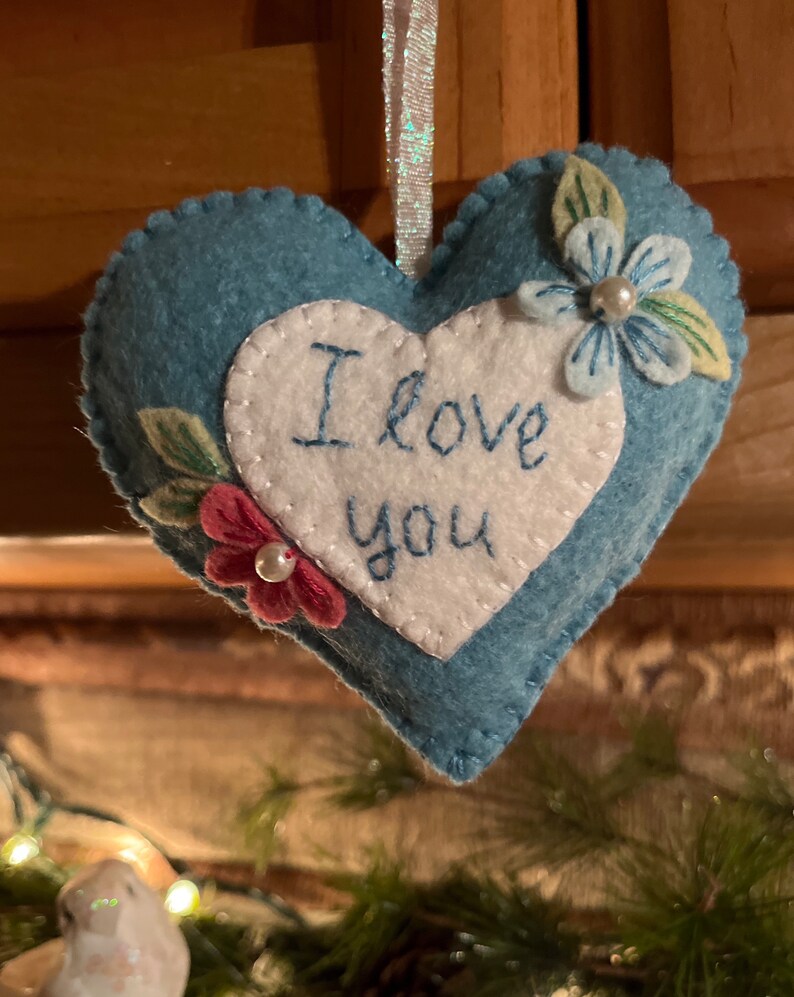 Może przedstawiać: Niebieska filcowa ozdoba w kształcie serca z białym sercem pośrodku. Na białym sercu wyhaftowano niebieskimi literami napis "I love you". Ozdoba jest ozdobiona filcowymi kwiatami i liśćmi i ma wstążkę do zawieszenia.