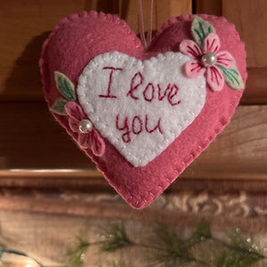 Może przedstawiać: R&oacute;żowa filcowa ozdoba w kształcie serca z wyhaftowanymi słowami "I love you" na białym sercu. Ozdoba jest ozdobiona r&oacute;żowymi kwiatami, zielonymi liśćmi i perłowymi akcentami. Do zawieszenia przymocowany jest r&oacute;żowy sznurek.