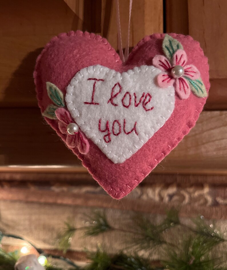 Może przedstawiać: R&oacute;żowa filcowa ozdoba w kształcie serca z wyhaftowanymi słowami "I love you" na białym sercu. Ozdoba jest ozdobiona r&oacute;żowymi kwiatami, zielonymi liśćmi i perłowymi akcentami. Do zawieszenia przymocowany jest r&oacute;żowy sznurek.