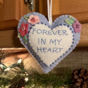 May include: A heart-shaped, light blue felt ornament with the words "FOREVER IN MY HEART" embroidered in blue. The ornament is decorated with pink and purple felt flowers and blue bead accents. A white ribbon is attached for hanging.