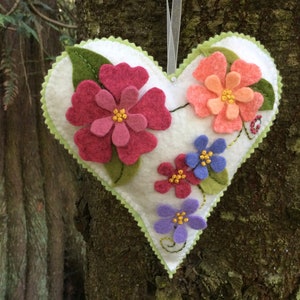May include: A heart-shaped felt decoration with a white base, adorned with colorful felt flowers in shades of pink, orange, purple, and blue. The edges are trimmed with green, and a white ribbon is attached for hanging. The decoration is displayed against a natural tree trunk background.