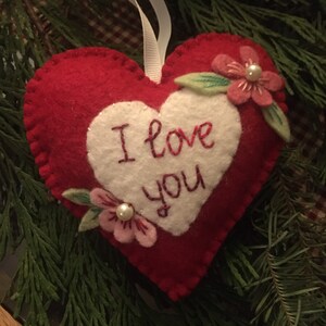 Może przedstawiać: Czerwona filcowa ozdoba w kształcie serca z białym sercem pośrodku. Słowa "I love you" są wyhaftowane na czerwono. R&oacute;żowe kwiaty z perłami i zielonymi liśćmi zdobią ozdobę. Do zawieszenia przymocowana jest biała wstążka.