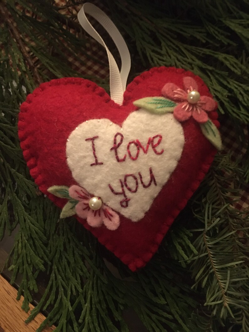 Może przedstawiać: Czerwona filcowa ozdoba w kształcie serca z białym sercem pośrodku. Słowa "I love you" są wyhaftowane na czerwono. R&oacute;żowe kwiaty z perłami i zielonymi liśćmi zdobią ozdobę. Do zawieszenia przymocowana jest biała wstążka.
