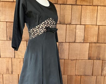 May include: A black dress with a scoop neckline and three-quarter sleeves. The waist features a decorative panel with a geometric pattern and a small bow. The dress has a flared skirt and is displayed on a mannequin.
