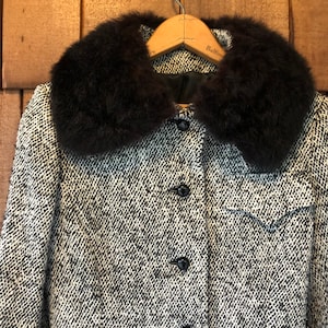 May include: A black and white tweed jacket with a large, black faux fur collar. The jacket has a button closure and a pocket on the left side.