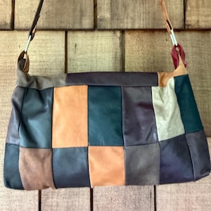 May include: A rectangular shoulder bag constructed from a patchwork of leather panels in various colors including dark brown, tan, forest green, and cream. The bag features a thin leather strap attached with silver-toned metal hardware rings.