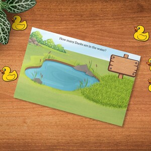 May include: A children's educational card with a pond scene and the text "How many Ducks are in the water?" is surrounded by yellow duck cutouts on a wooden surface. A green plant is in the upper left corner.