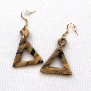 May include: A pair of gold triangle earrings with a marbled pattern in shades of brown and black. The earrings have a round, gold accent at the top.