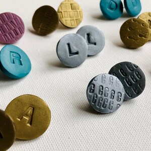 Könnte beinhalten: Eine Sammlung runder, bunter Tonohrringe mit verschiedenen Mustern und Buchstaben. Die Ohrringe sind auf einer weißen Oberfläche angeordnet.