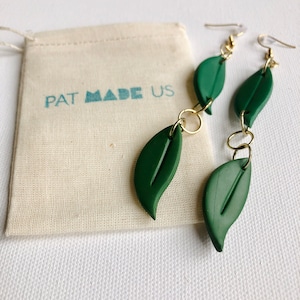 May include: A pair of green leaf dangle earrings with gold chain accents. The earrings are packaged in a small white fabric bag with the text "PAT MADE US" printed on it.