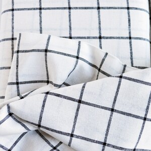 Pu&ograve; includere: Primo piano di tessuto bianco con motivo a griglia nera. Il tessuto &egrave; piegato, mostrando la trama e il design. La griglia &egrave; composta da linee nere distanziate uniformemente, creando un look classico e minimalista. Il tessuto sembra essere lino o un materiale simile.