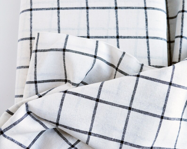 Pu&ograve; includere: Primo piano di tessuto bianco con motivo a griglia nera. Il tessuto &egrave; piegato, mostrando la trama e il design. La griglia &egrave; composta da linee nere distanziate uniformemente, creando un look classico e minimalista. Il tessuto sembra essere lino o un materiale simile.