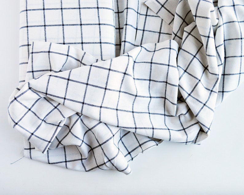 Pu&ograve; includere: Tessuto bianco con motivo a griglia blu navy. Il tessuto &egrave; piegato e drappeggiato, mostrando la trama e il design. Il motivo &egrave; un classico a quadri, adatto a vari progetti di arredamento o abbigliamento.