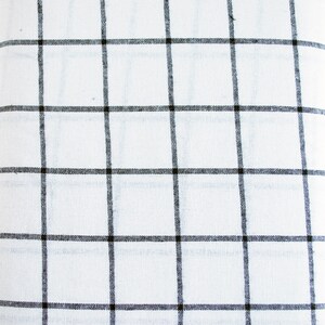 Pu&ograve; includere: Un tessuto bianco arrotolato con un motivo a griglia nera. Le linee della griglia sono uniformemente distanziate, creando un design a scacchi. Il tessuto sembra essere un materiale leggero, forse lino o cotone.