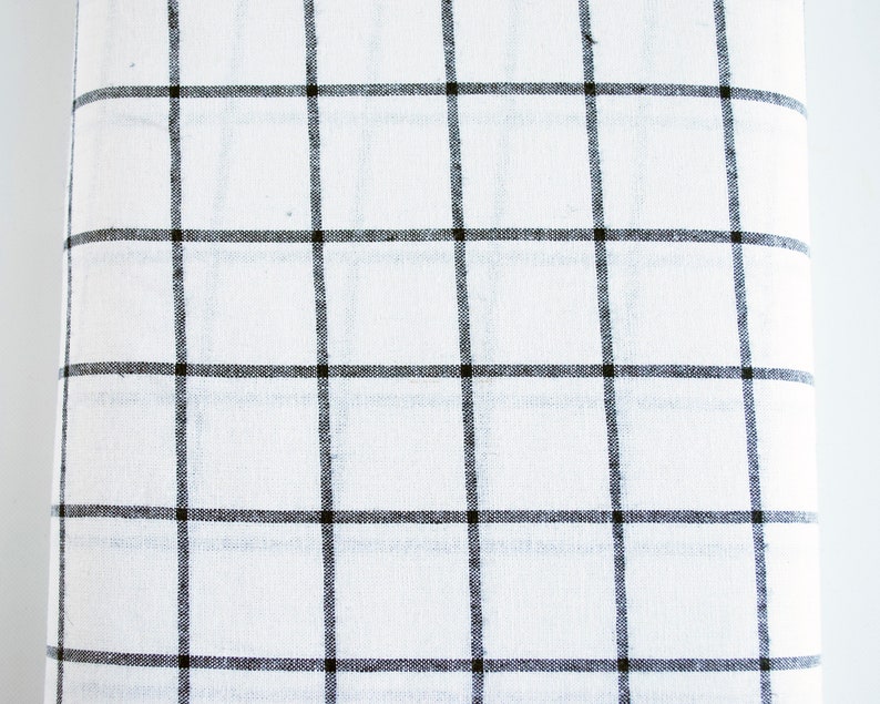 Pu&ograve; includere: Un tessuto bianco arrotolato con un motivo a griglia nera. Le linee della griglia sono uniformemente distanziate, creando un design a scacchi. Il tessuto sembra essere un materiale leggero, forse lino o cotone.