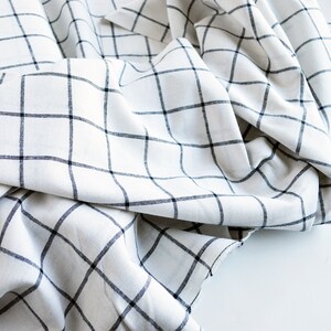 Pu&ograve; includere: Tessuto bianco con un motivo a griglia nera. Il tessuto &egrave; drappeggiato e piegato, creando ombre e consistenza. Le linee della griglia sono uniformemente distanziate, formando un classico motivo a quadri. Il tessuto sembra essere un materiale leggero.