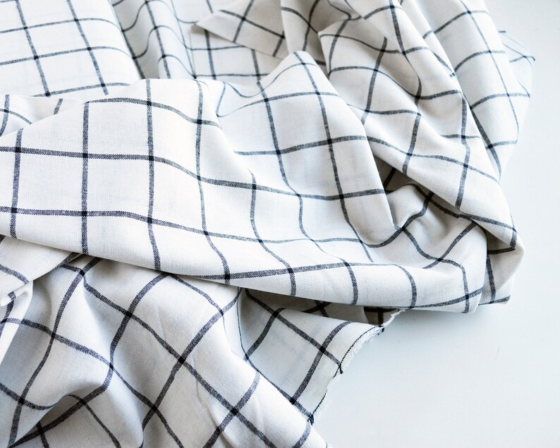 Pu&ograve; includere: Tessuto bianco con un motivo a griglia nera. Il tessuto &egrave; drappeggiato e piegato, creando ombre e consistenza. Le linee della griglia sono uniformemente distanziate, formando un classico motivo a quadri. Il tessuto sembra essere un materiale leggero.