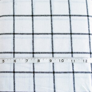 Pu&ograve; includere: Tessuto bianco con motivo a griglia nera. Un metro a nastro &egrave; posizionato sul tessuto, mostrando misure da 10 a 33 cm. Adatto per cucito o progetti artigianali.