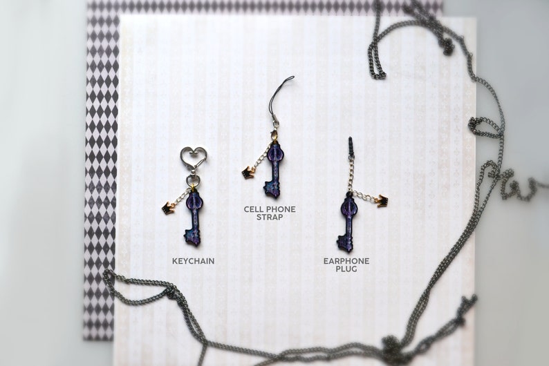 May include: Three key-shaped charms with gold accents and a purple and blue design. The charms are attached to different chains: a keychain, a cell phone strap, and an earphone plug.