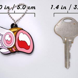 May include: A colorful keychain charm, approximately 2.0 inches (5.0 cm) long, featuring a cartoon design with pink, white, and brown elements. A silver key, about 1.4 inches (3.5 cm) long, is next to the charm. Both objects are on a white background.