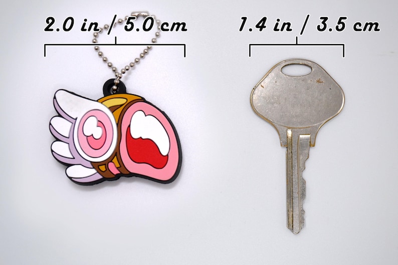 May include: A colorful keychain charm, approximately 2.0 inches (5.0 cm) long, featuring a cartoon design with pink, white, and brown elements. A silver key, about 1.4 inches (3.5 cm) long, is next to the charm. Both objects are on a white background.