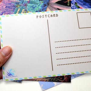 Pode incluir: Postal branco com a palavra "POSTCARD" impressa no topo. O postal tem uma borda azul e verde e uma linha vertical marrom para o endereço. O postal é segurado em uma mão.