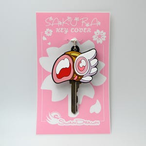 May include: A key cover with a cartoon design, featuring a pink, white, and red motif, and the text "SAKURA KEY COVER". The key cover is attached to a key and displayed on a pink card with a white flower design.
