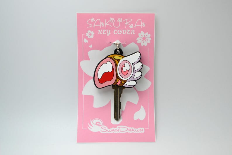 May include: A key cover with a cartoon design, featuring a pink, white, and red motif, and the text "SAKURA KEY COVER". The key cover is attached to a key and displayed on a pink card with a white flower design.