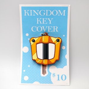 May include: A yellow and orange key cover with a white center and black accents. The key cover is shaped like a shield and has the text "KINGDOM KEY COVER" on a blue background with white polka dots. The price is $10.