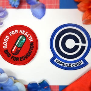 以下が含まれることがあります： 2つの刺繍ワッペン。1つは赤色で、薬のグラフィックと「GOOD FOR HEALTH BAD FOR EDUCATION」の文字が入っています。もう1つは青と白で、「C」のロゴと「CAPSULE CORP」の文字が入っています。装飾的な花がワッペンを囲んでいます。