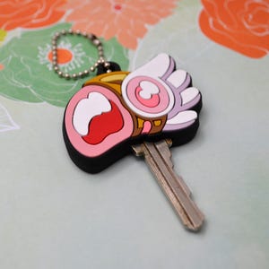 May include: A key with a decorative charm. The charm is a cartoon-style design with a black border, pink, white, red, and gold accents. The key is silver and attached to a silver ball chain.