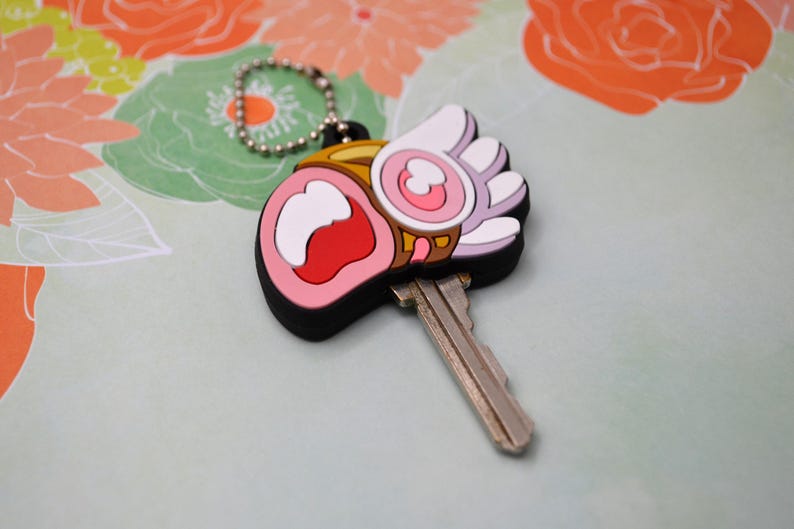 May include: A key with a decorative charm. The charm is a cartoon-style design with a black border, pink, white, red, and gold accents. The key is silver and attached to a silver ball chain.