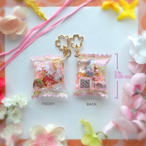 May include: Two clear, 3-inch (7.62 cm) tall, candy-shaped keychains with gold-colored flower-shaped clasps. One keychain shows a cartoon character in a red dress, the other shows a cartoon character in a purple dress. Both are filled with pink confetti.