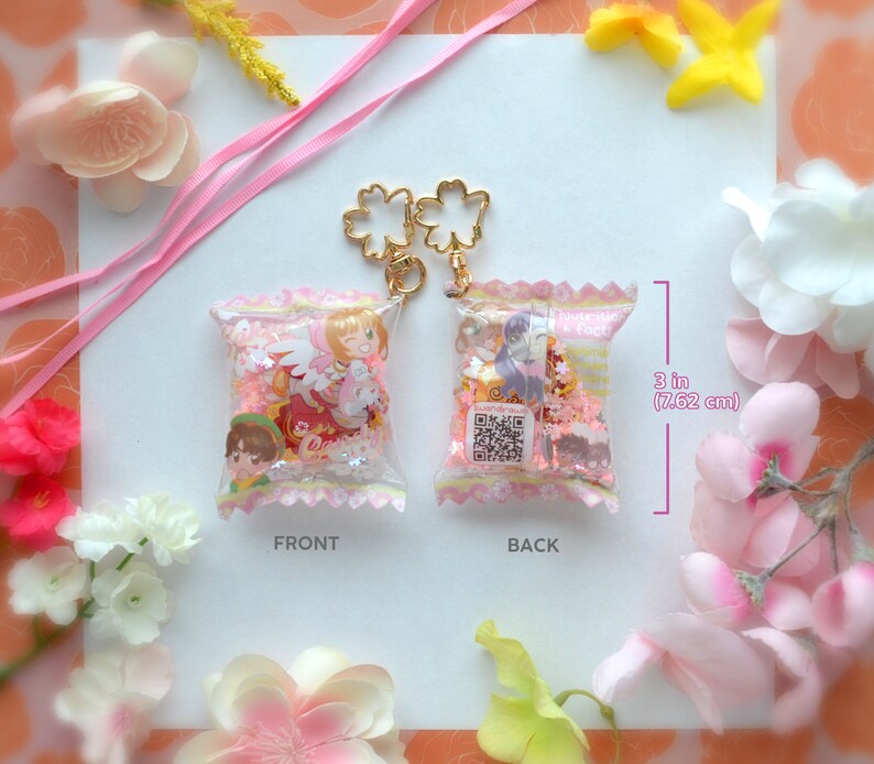May include: Two clear, 3-inch (7.62 cm) tall, candy-shaped keychains with gold-colored flower-shaped clasps. One keychain shows a cartoon character in a red dress, the other shows a cartoon character in a purple dress. Both are filled with pink confetti.