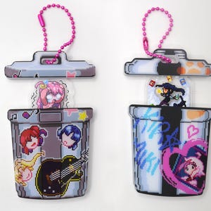 Op de afbeelding: Twee sleutelhangers in pixel art-stijl. Eén heeft een vuilnisbakontwerp met anime-personages en een gitaar. De andere heeft een vergelijkbaar vuilnisbakontwerp met anime-personages en de tekst "MIKI". Beide hebben roze kralenkettingen.