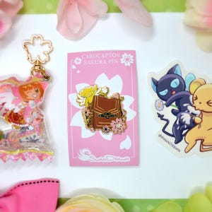 May include: A collection of anime-themed merchandise, including a clear keychain with character figures, a pink pin featuring a brown backpack design, and a sticker with two cartoon characters. The items are arranged on a white surface with pink floral accents.