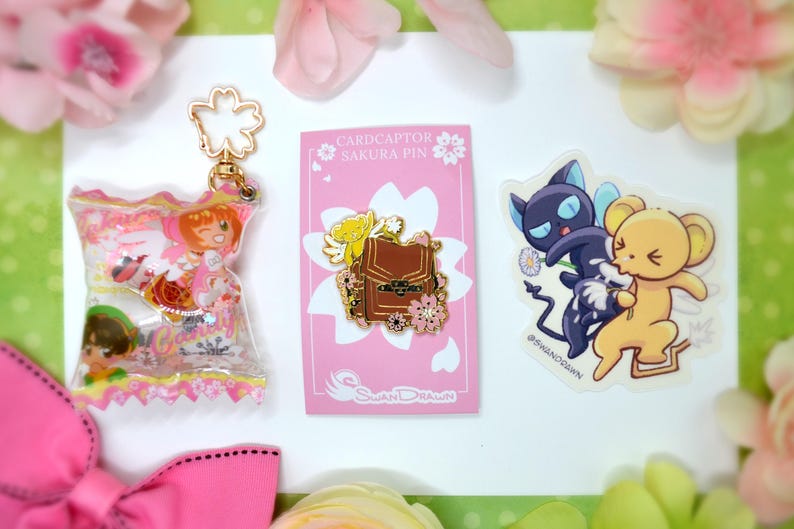 May include: A collection of anime-themed merchandise, including a clear keychain with character figures, a pink pin featuring a brown backpack design, and a sticker with two cartoon characters. The items are arranged on a white surface with pink floral accents.