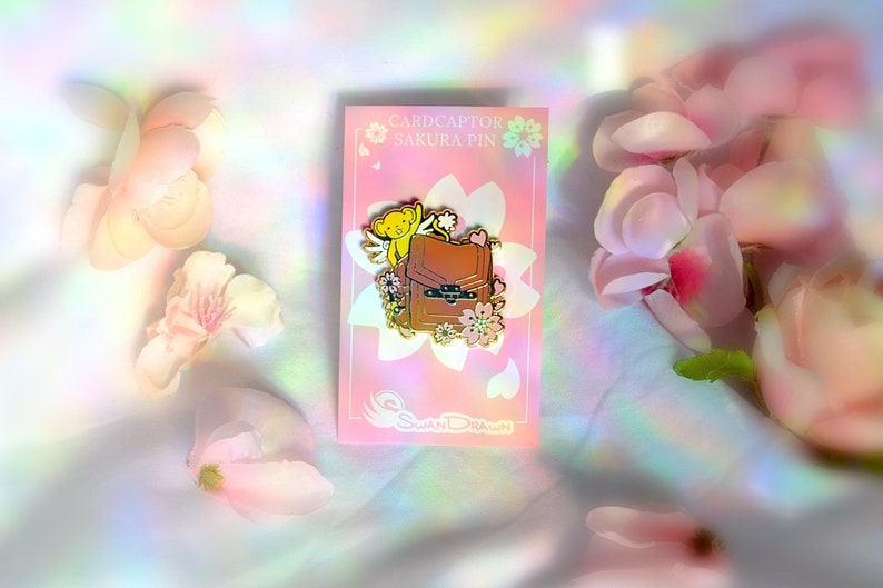May include: A pink card with a pin featuring a brown backpack, a yellow creature with wings, and pink flowers. The card reads "Cardcaptor Sakura Pin" and "Swan Drawn". Pink flowers are scattered around the card.