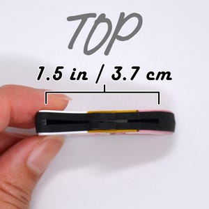 May include: A small, rectangular object with a black center, white and pink edges, and a gold accent. The text "TOP" is above the object, and the dimensions are 1.5 inches / 3.7 cm.