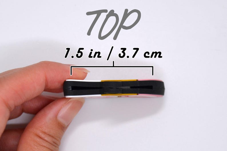 May include: A small, rectangular object with a black center, white and pink edges, and a gold accent. The text "TOP" is above the object, and the dimensions are 1.5 inches / 3.7 cm.