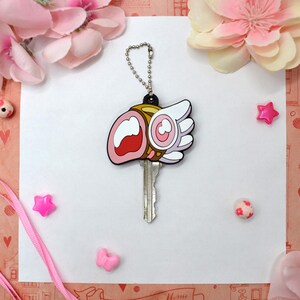 May include: A key with a decorative cover featuring a cartoon design with pink, white, and gold accents. The key is attached to a silver chain. The background includes pink flowers and decorative elements.