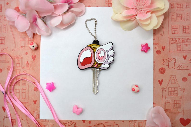 May include: A key with a decorative cover featuring a cartoon design with pink, white, and gold accents. The key is attached to a silver chain. The background includes pink flowers and decorative elements.
