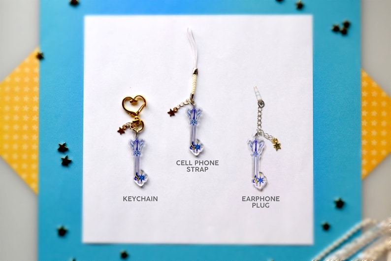 May include: Three keychains featuring a blue and white keyblade design. The keychains are displayed on a white background with the words "KEYCHAIN", "CELL PHONE STRAP", and "EARPHONE PLUG" printed below each keychain.
