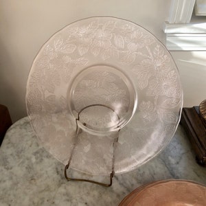 Puede incluir: Un plato de servir de vidrio transparente con un diseño floral grabado. El plato está en un soporte de metal.