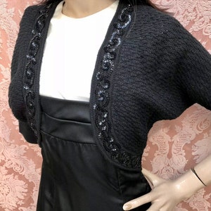 May include: Black knitted shrug with a beaded trim. The trim features a swirling design.