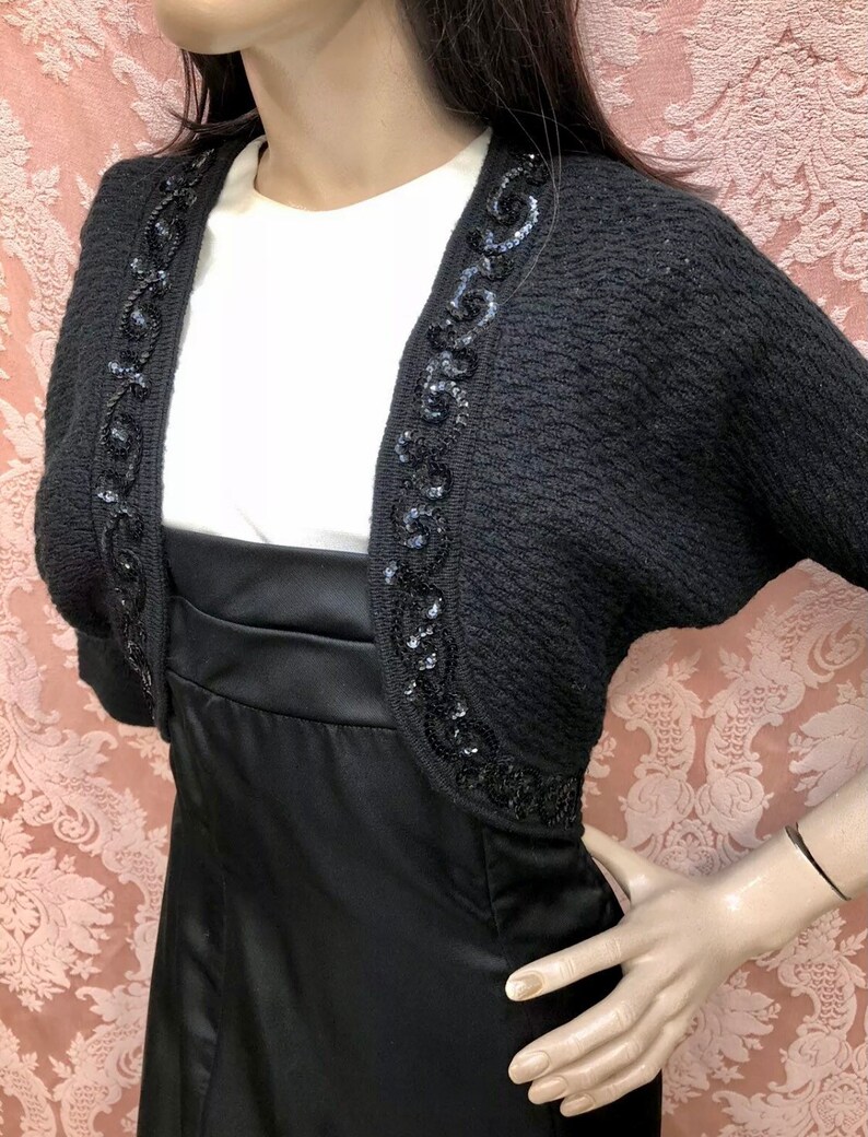 May include: Black knitted shrug with a beaded trim. The trim features a swirling design.